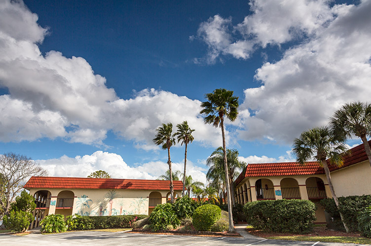 Exterior shot of our family friendly resort in Kissimmee, FL. Red tile roofing, and large palm trees surrounding an empty parking lot